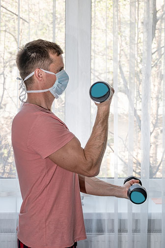 man lifting dumbbells at home while wearing a protective mask